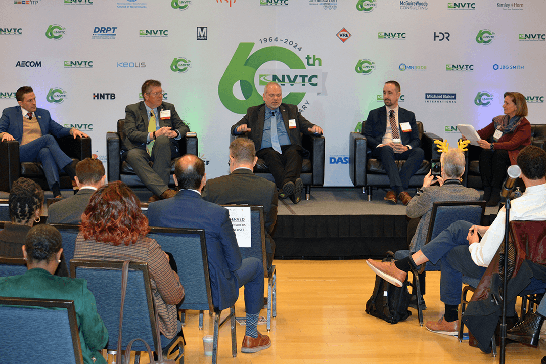 Four men in suits sitting in chairs on a stage with one woman in chair. Seated in front of them listening.