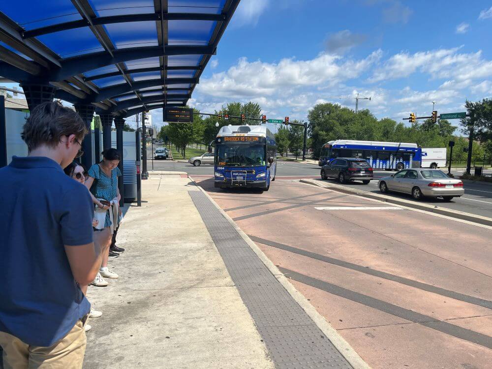 Bus passengers waiting under the shade of a blue bus awning while bus pulls in to a red bus only lane next to the bus stop.