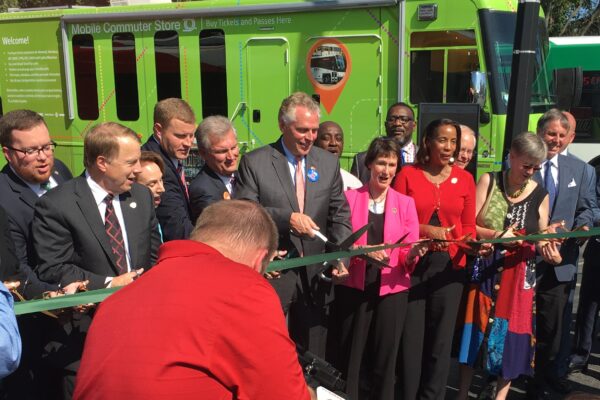 Group of men and women in suits, holding green ribbon. VA Governor McAuliffe with giant scissors at center.
