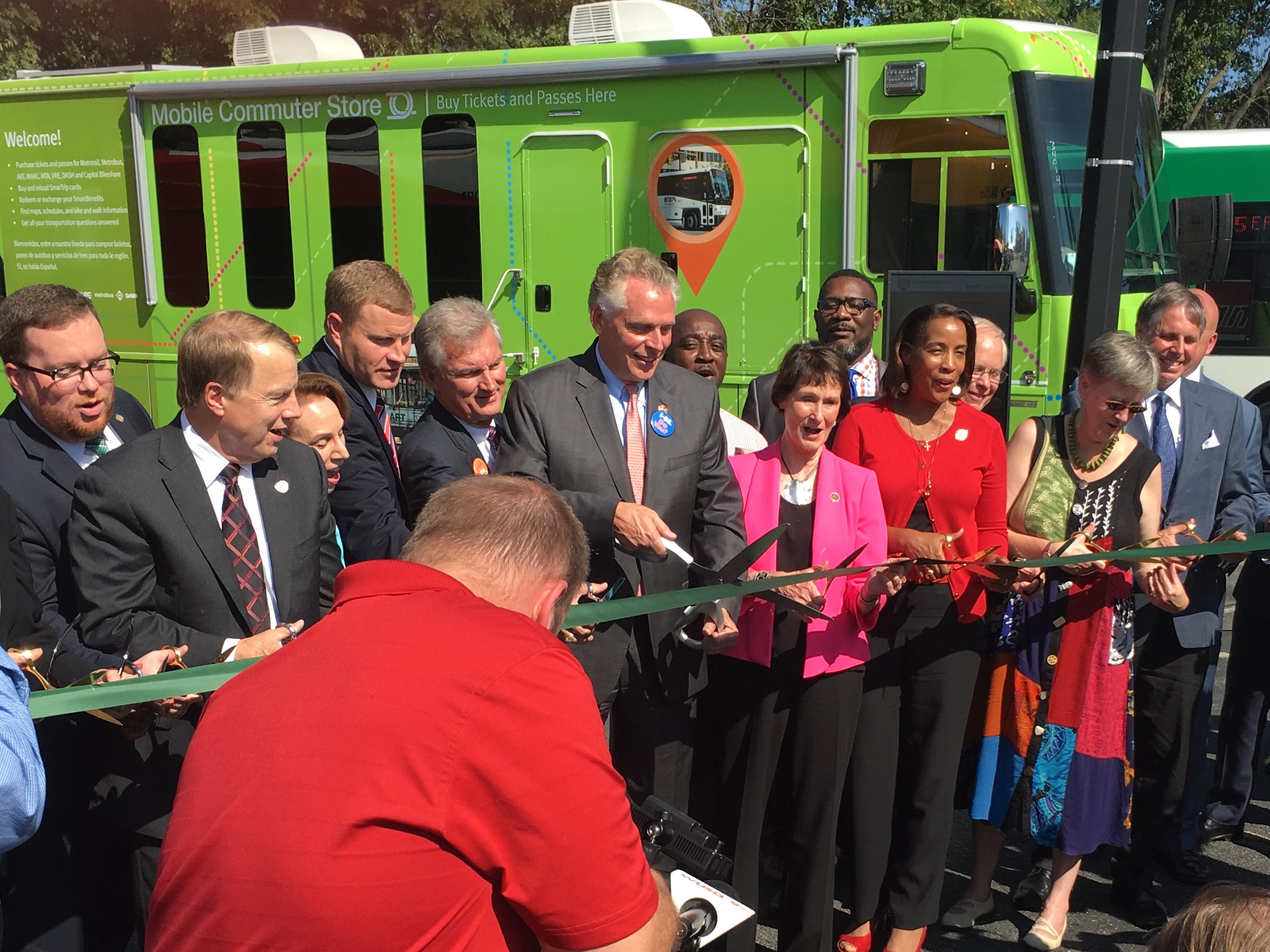 Group of men and women in suits, holding green ribbon. VA Governor McAuliffe with giant scissors at center.